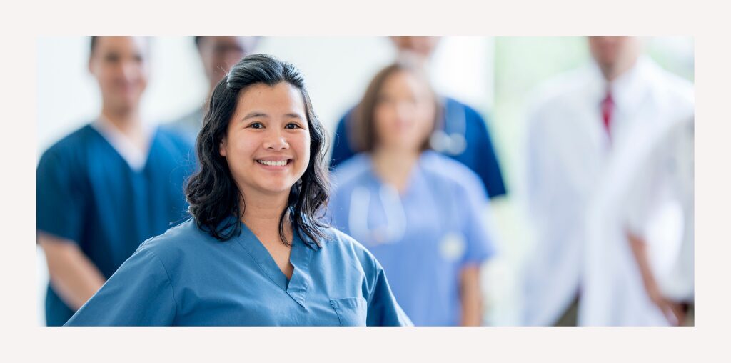A youthful nurse in blue scrubs stands confidently in the foreground. Her medical colleagues stand out of focus in the background.