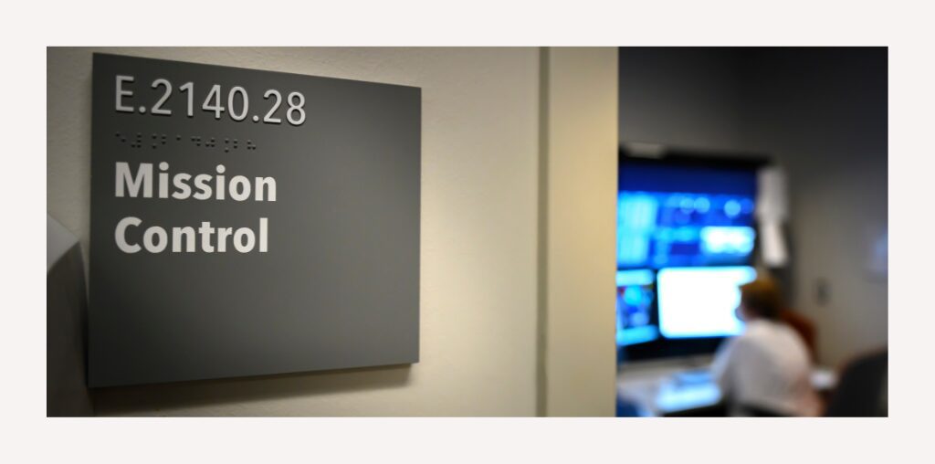 The nameplate identifying the entryway of Texas Children's Mission Control. An out-of-focus employee sits at a panel of monitors in the background.